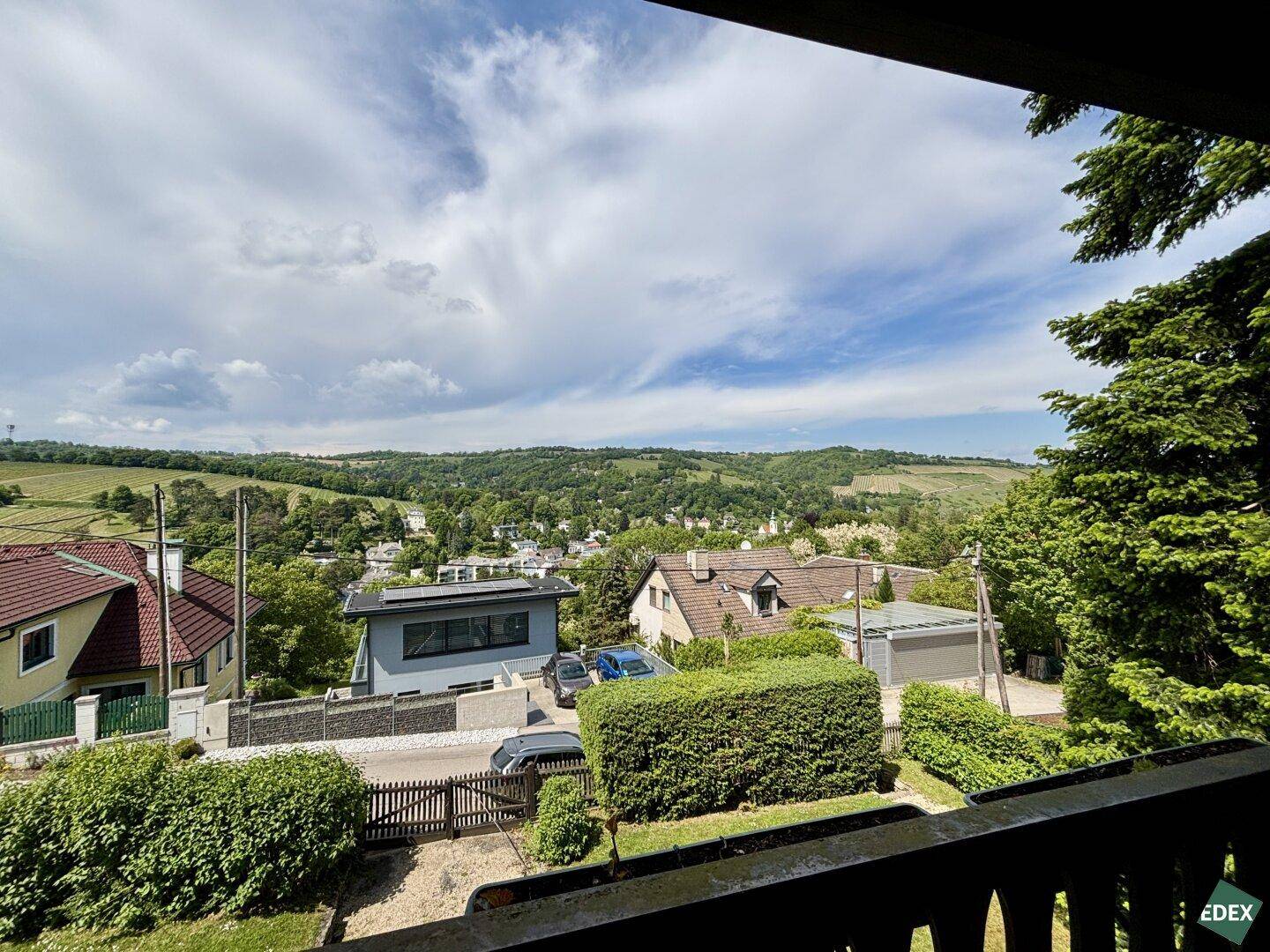 Aussicht von einem Balkon auf die umliegende Wohngegend und grüne Hügel unter Wolken.