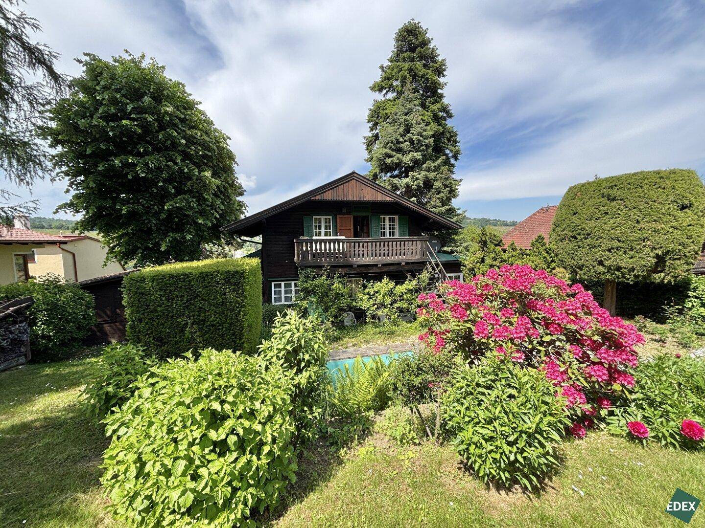 Traditionelles Holzhaus umgeben von einem blühenden Garten mit Sträuchern und Bäumen.