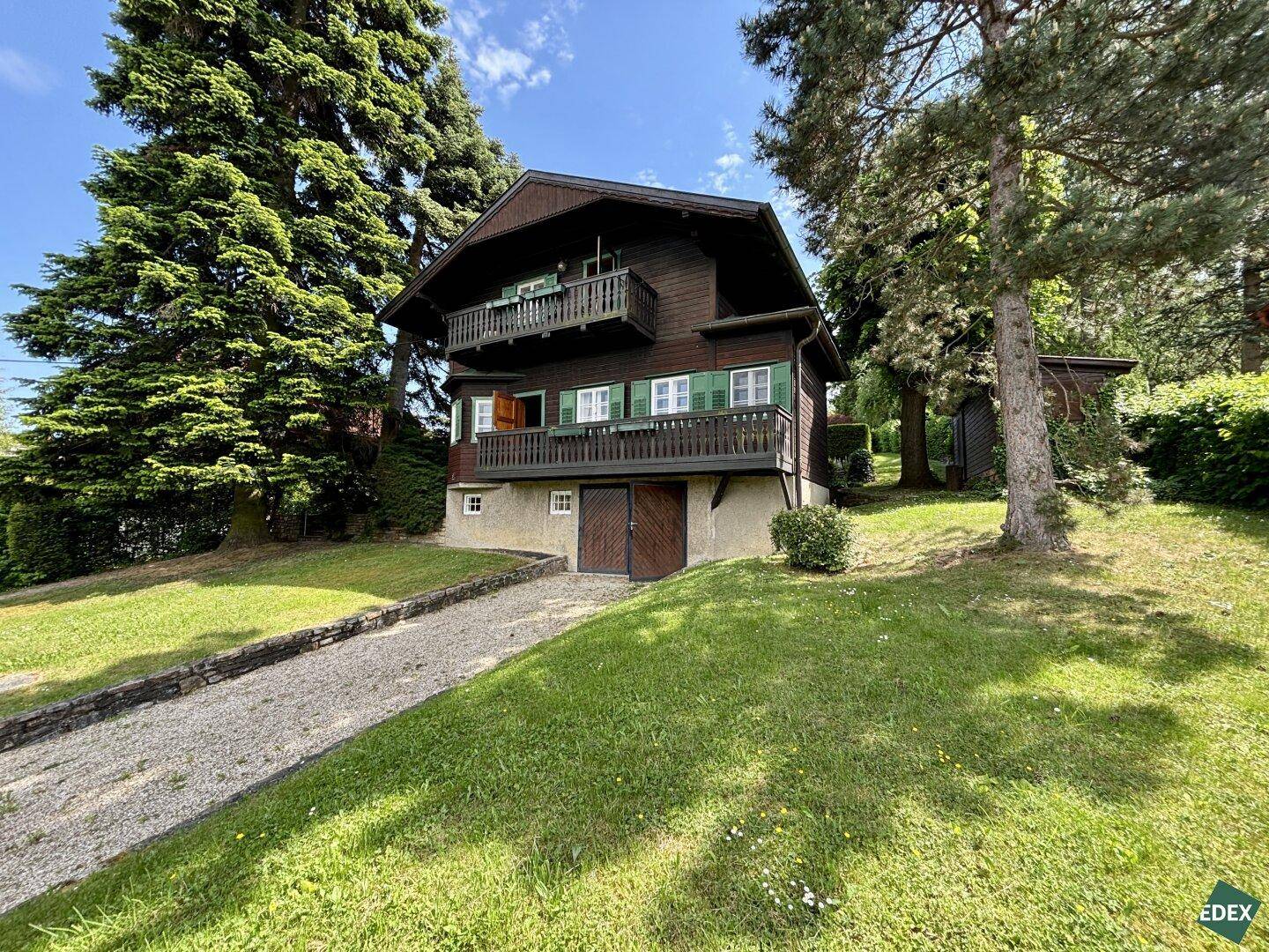 Holzhaus im Chalet-Stil mit Kiesauffahrt und umgebender grüner Landschaft.