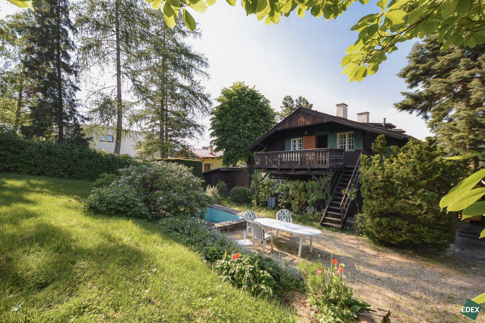 Gartenbereich mit Kiesweg, Sitzgelegenheiten und einem kleinen Pool, umgeben von blühenden Pflanzen und Bäumen.