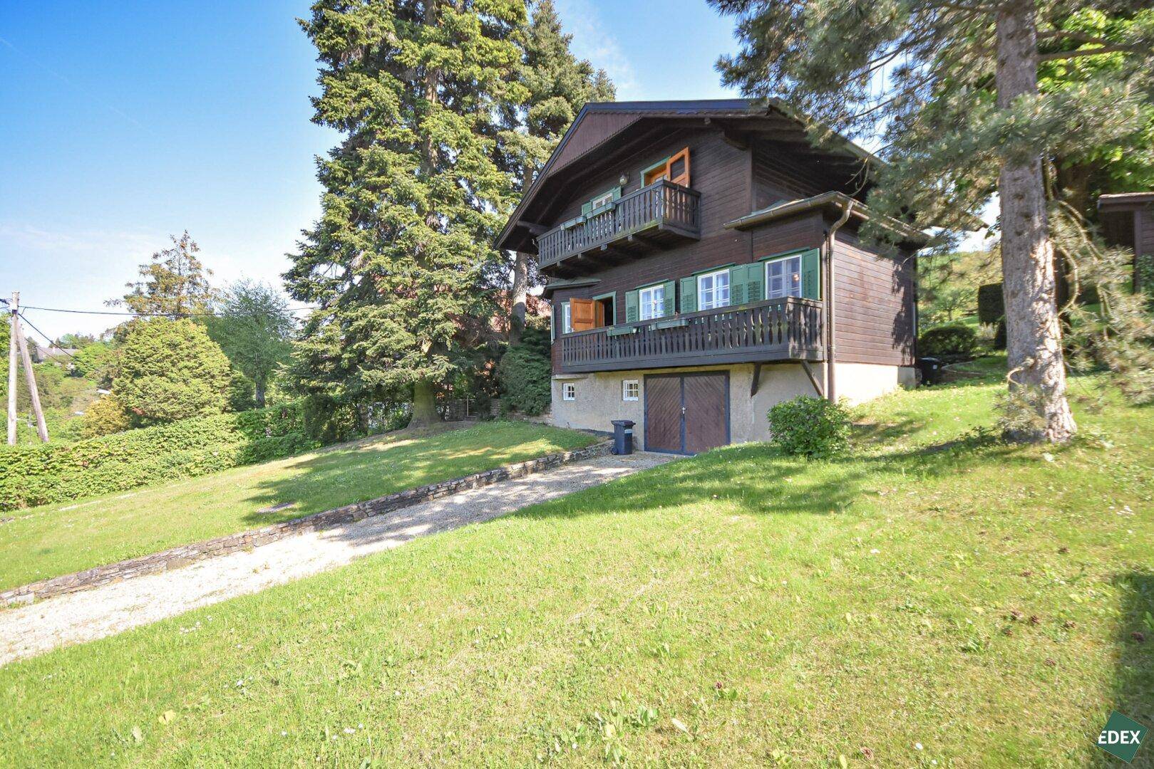 Traditionelles Holzhaus mit Balkonen und Zufahrt, umgeben von einem gepflegten Rasen und hohen Bäumen.