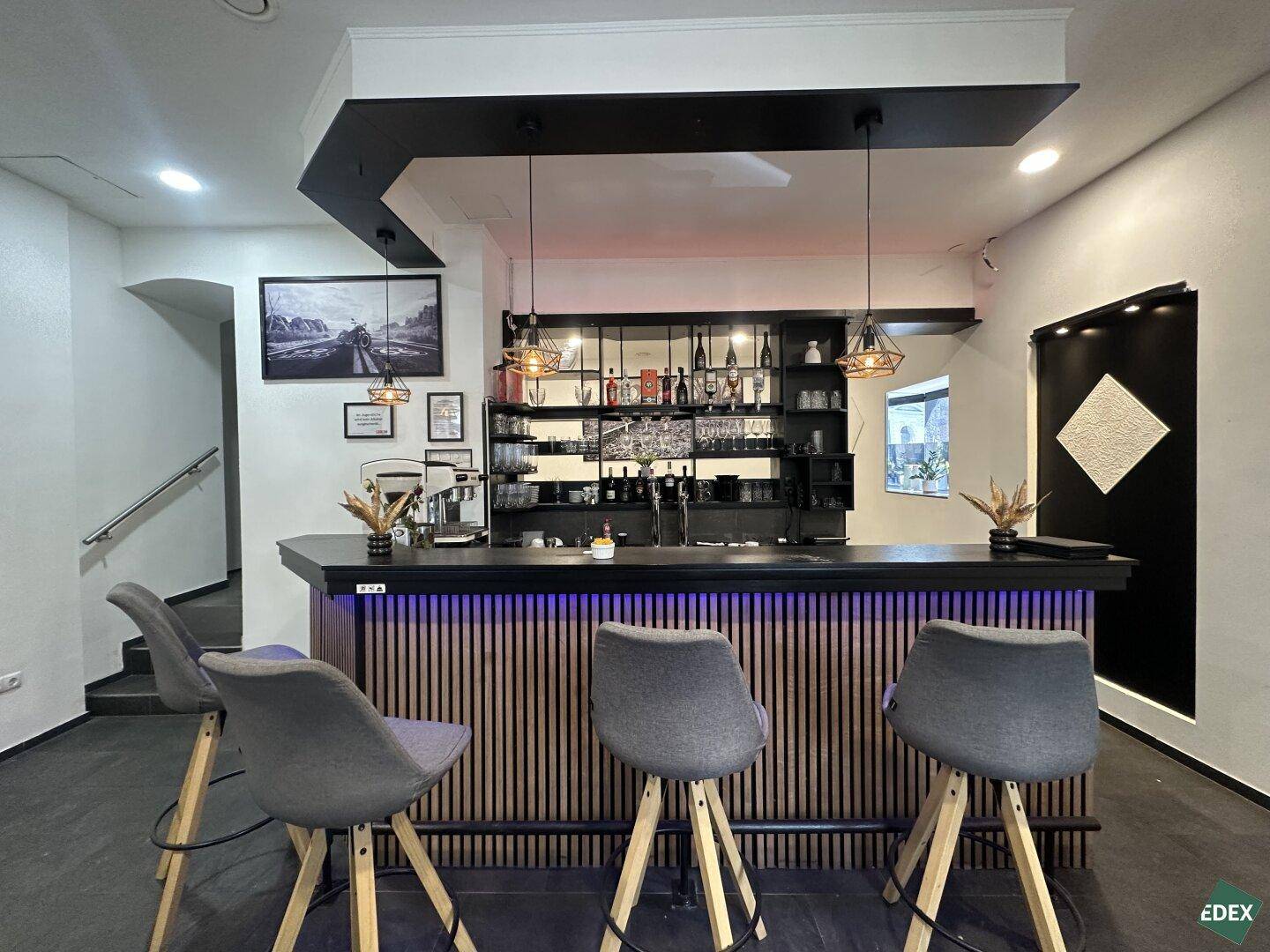 Moderne Bar mit Holzlamellenfront, dunkler Theke und stilvollen Barhockern in einem Innenraum.
