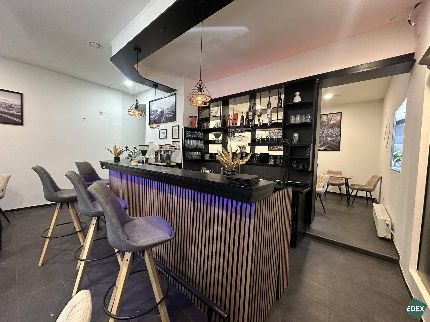Blick auf die moderne Bar mit Sitzgelegenheiten und einem Durchgang zu weiteren Tischen im Hintergrund.