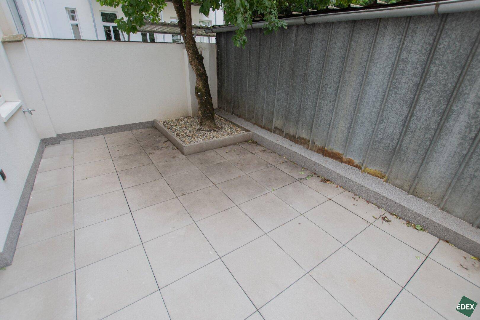 Ruhige Terrasse mit hellen Fliesen, einem Baum und einer hohen Abgrenzung, ideal zum Entspannen.