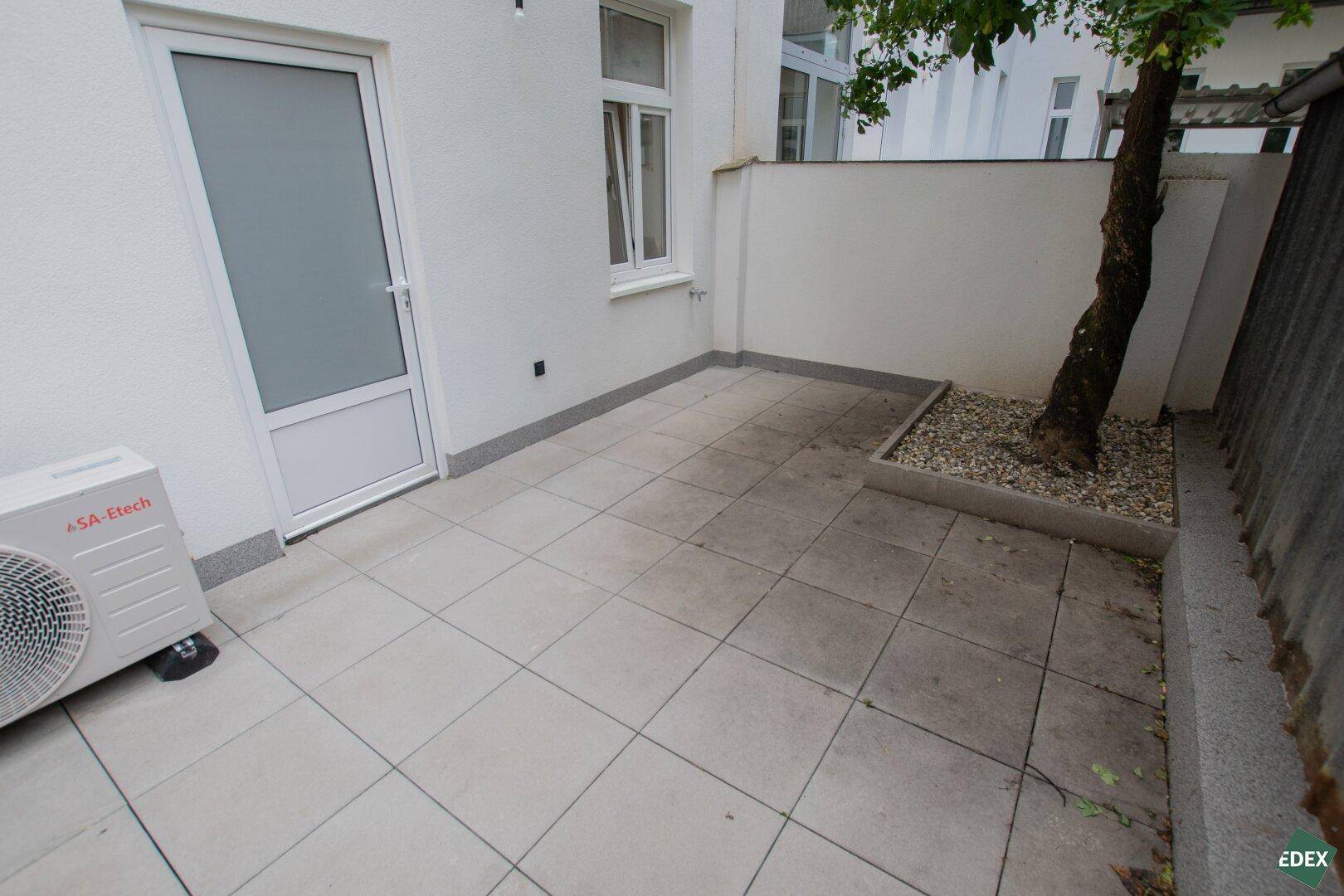Geflieste Terrasse mit einem kleinen Baum in einem Hochbeet und einer Klimaanlage-Außeneinheit.