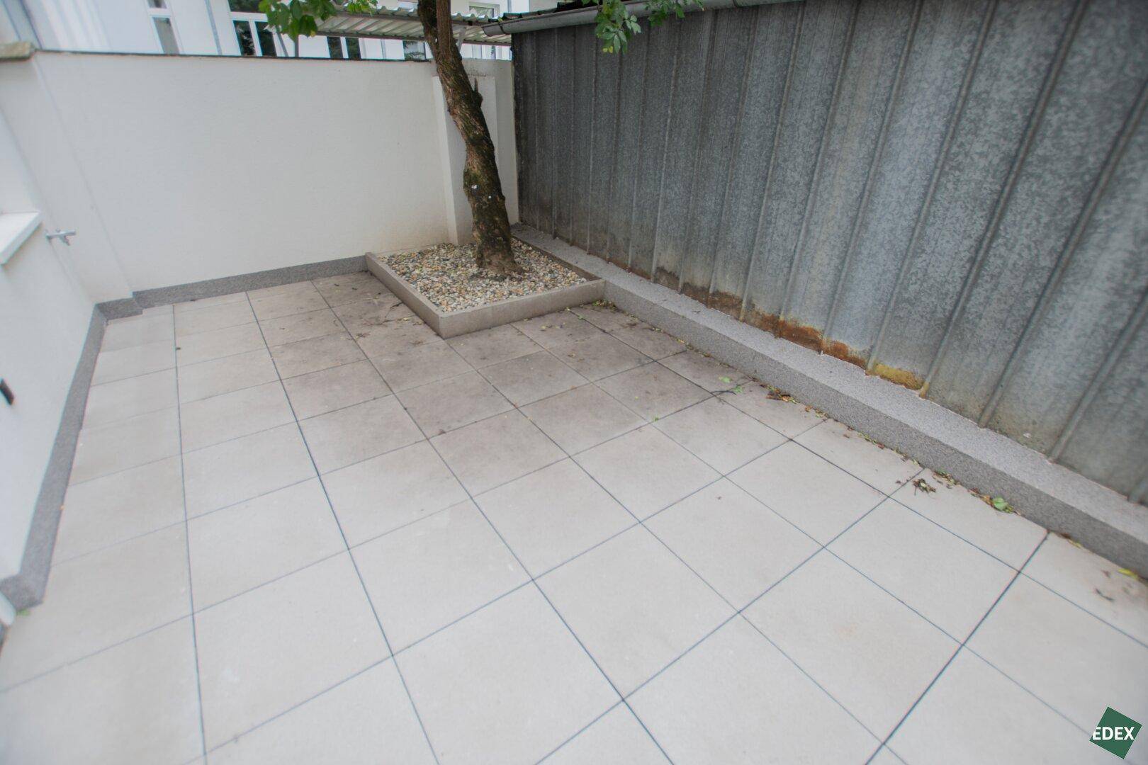 Gemütliche Terrasse mit hellen Bodenfliesen und einem kleinen Baum, umgeben von einer Mauer.