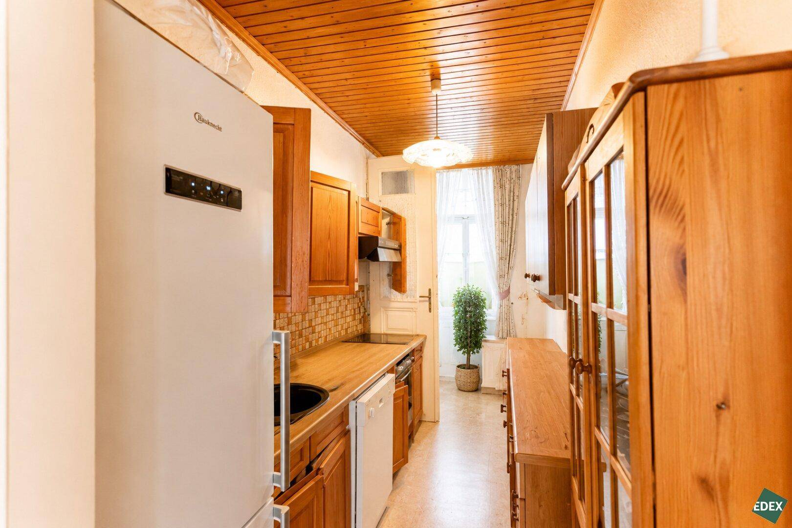 Funktionale Küche mit Holzschränken, Kühlschrank und Blick auf einen kleinen Baum im Fenster.