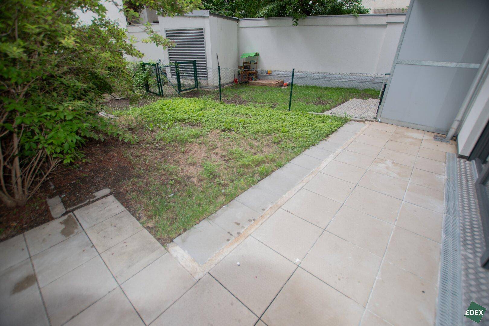 Gepflasterte Terrasse mit angrenzendem kleinen Gartenbereich und Grünflächen.