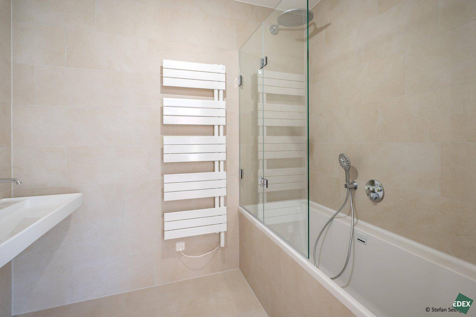 Modernes Badezimmer mit Badewanne, Glasduschwand, beheiztem Handtuchhalter und beigefarbenen Fliesen.