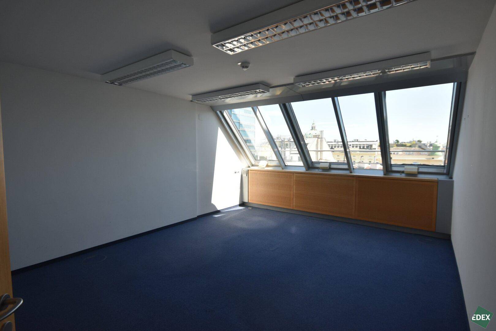 Leeres Büro mit blauem Teppichboden, schrägen Fenstern und Blick auf die Stadtlandschaft.