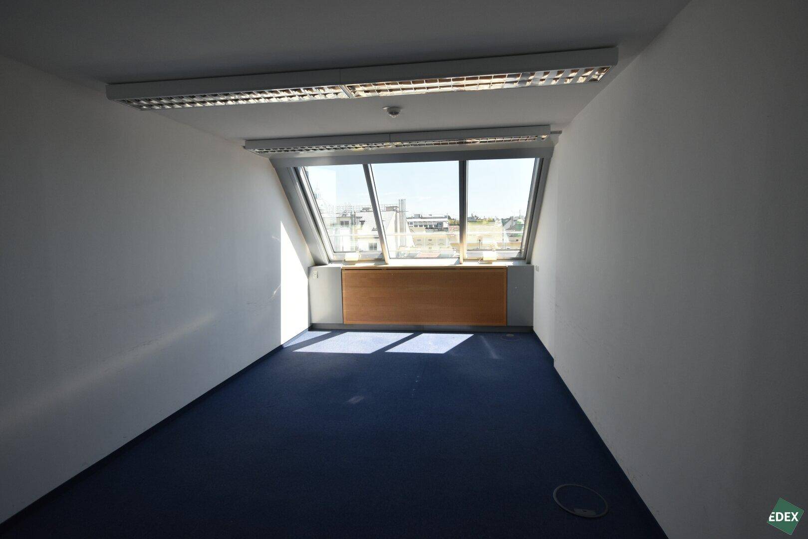 Helles Büro mit blauem Teppichboden und großen, schrägen Fenstern, die einen Stadtblick bieten.