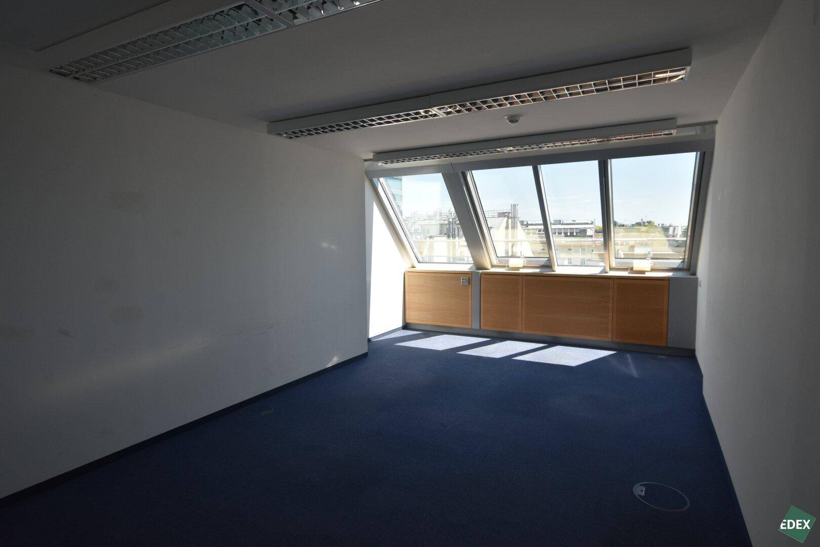 Geräumiges Büro mit viel Tageslicht durch die markanten, schrägen Fenster und blauen Teppichboden.