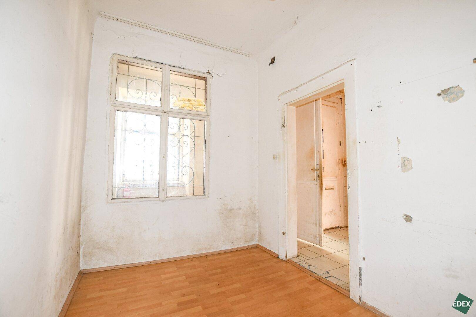 Leerer Raum mit Fenster und Blick auf eine Tür, die zu einem gefliesten Bereich führt.