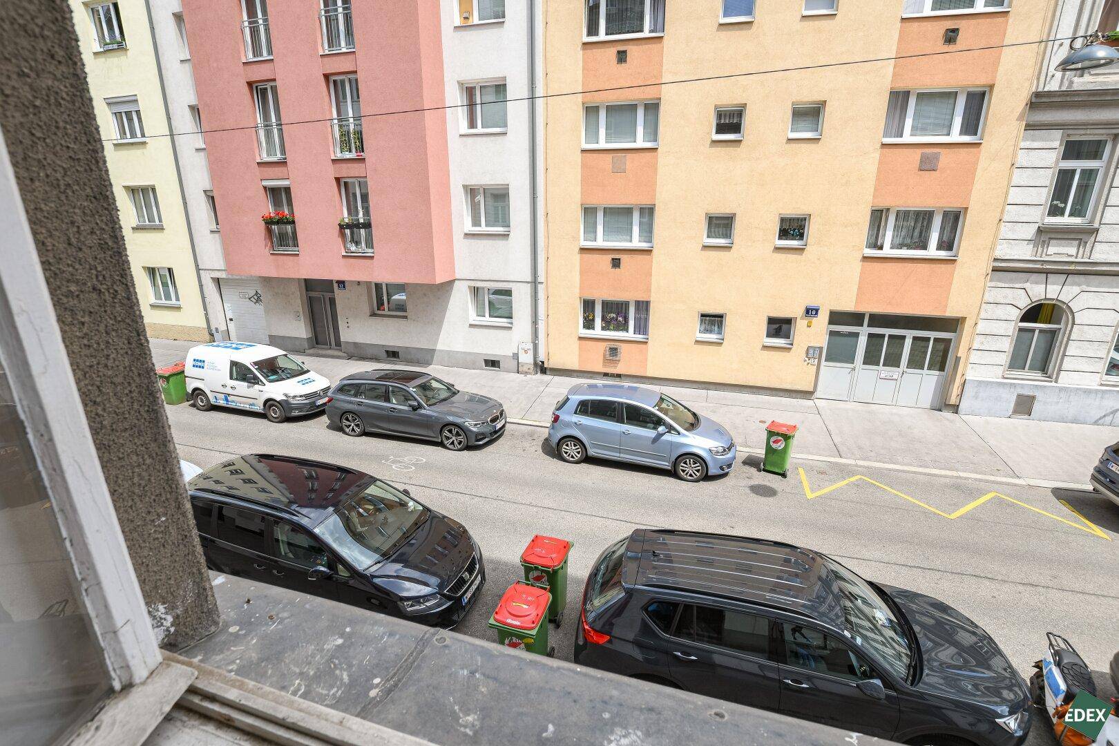 Blick aus dem Fenster auf eine belebte Straße mit parkenden Autos und bunten Gebäudefassaden.