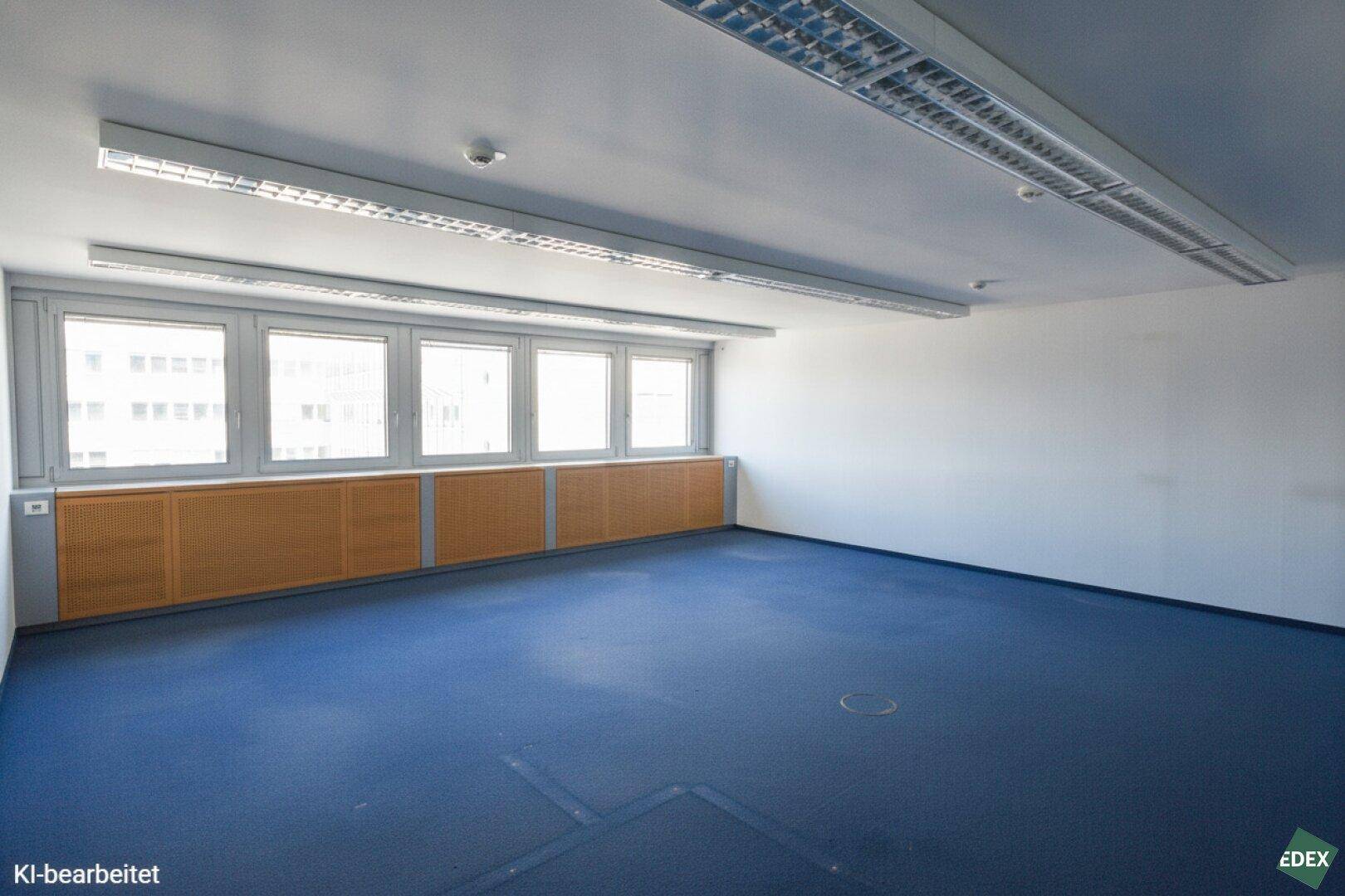 Leeres Büro mit blauem Teppichboden, großen Fenstern und integrierten Heizungsverkleidungen.