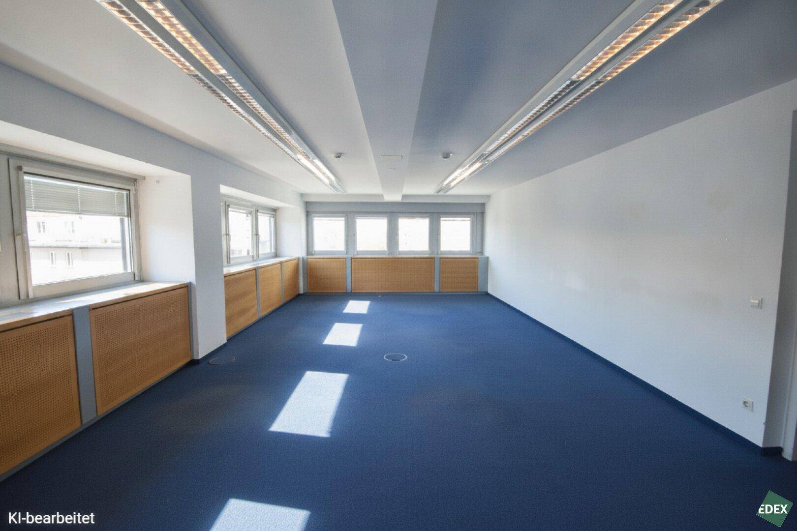 Geräumiges, leeres Büro mit blauem Teppichboden, vielen Fenstern und viel natürlichem Lichteinfall.