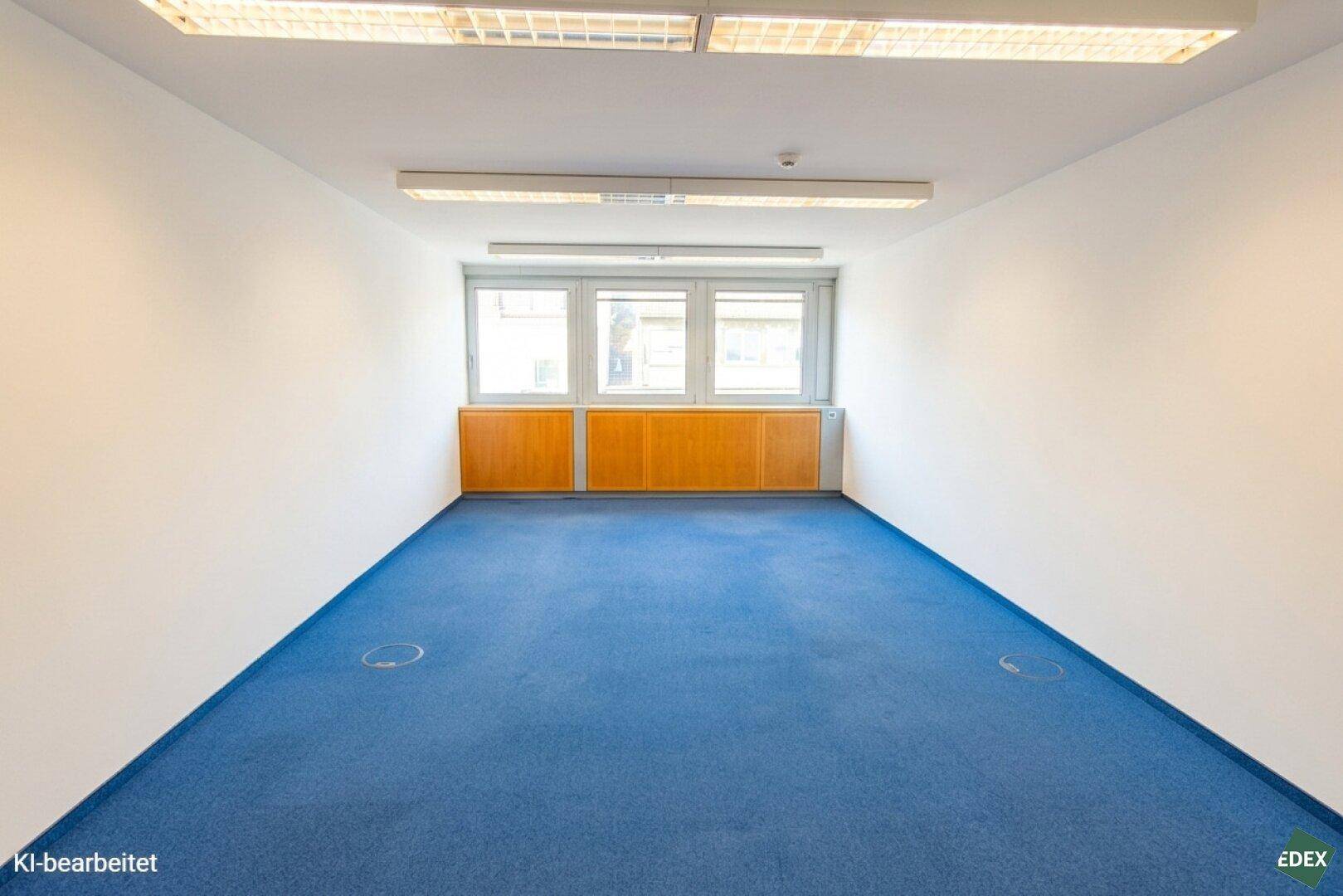 Helles, leeres Büro mit blauer Teppichauslage, rechteckigen Fenstern und Holzverkleidung unter den Fenstern.