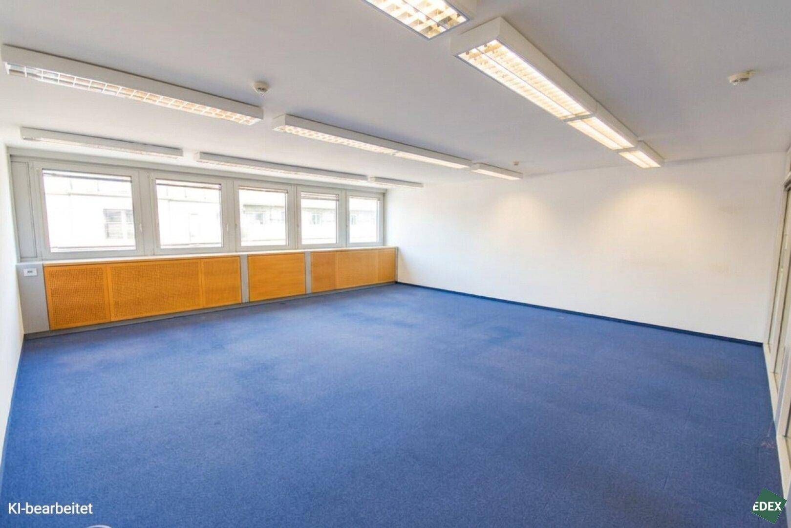Helles Büro mit blauem Teppichboden, großen Fenstern und funktionaler Deckenbeleuchtung.
