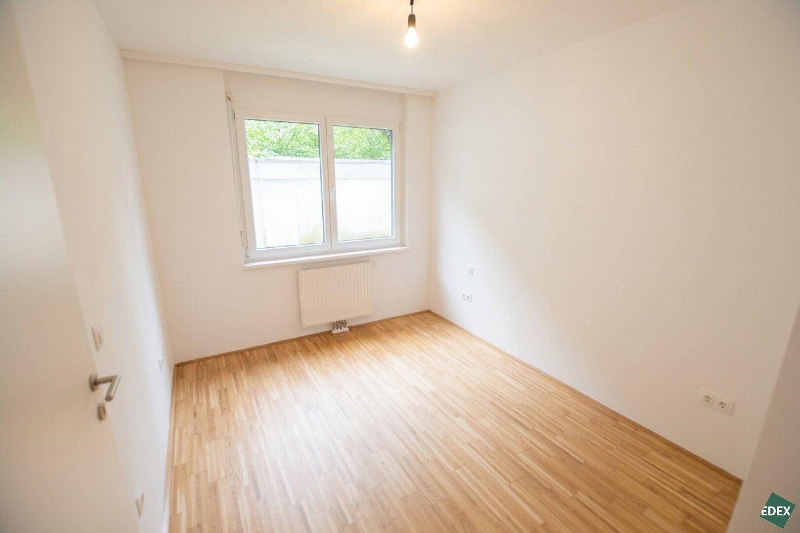 Helles Schlafzimmer mit Parkettboden und großem Fenster mit Blick ins Grüne.
