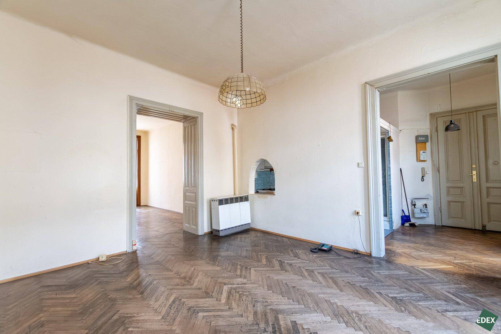 Geräumiges Zimmer mit Fischgrätparkett, Heizkörper und Durchgängen zu anderen Bereichen.