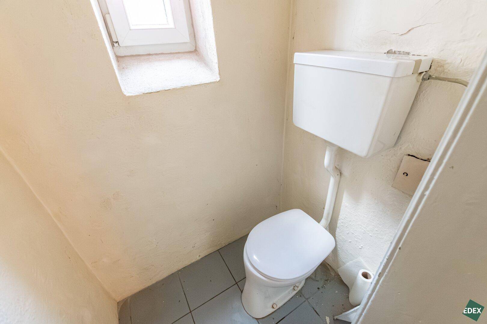 Einfaches WC mit Fenster, Fliesenboden und sichtbarem Spülkasten.