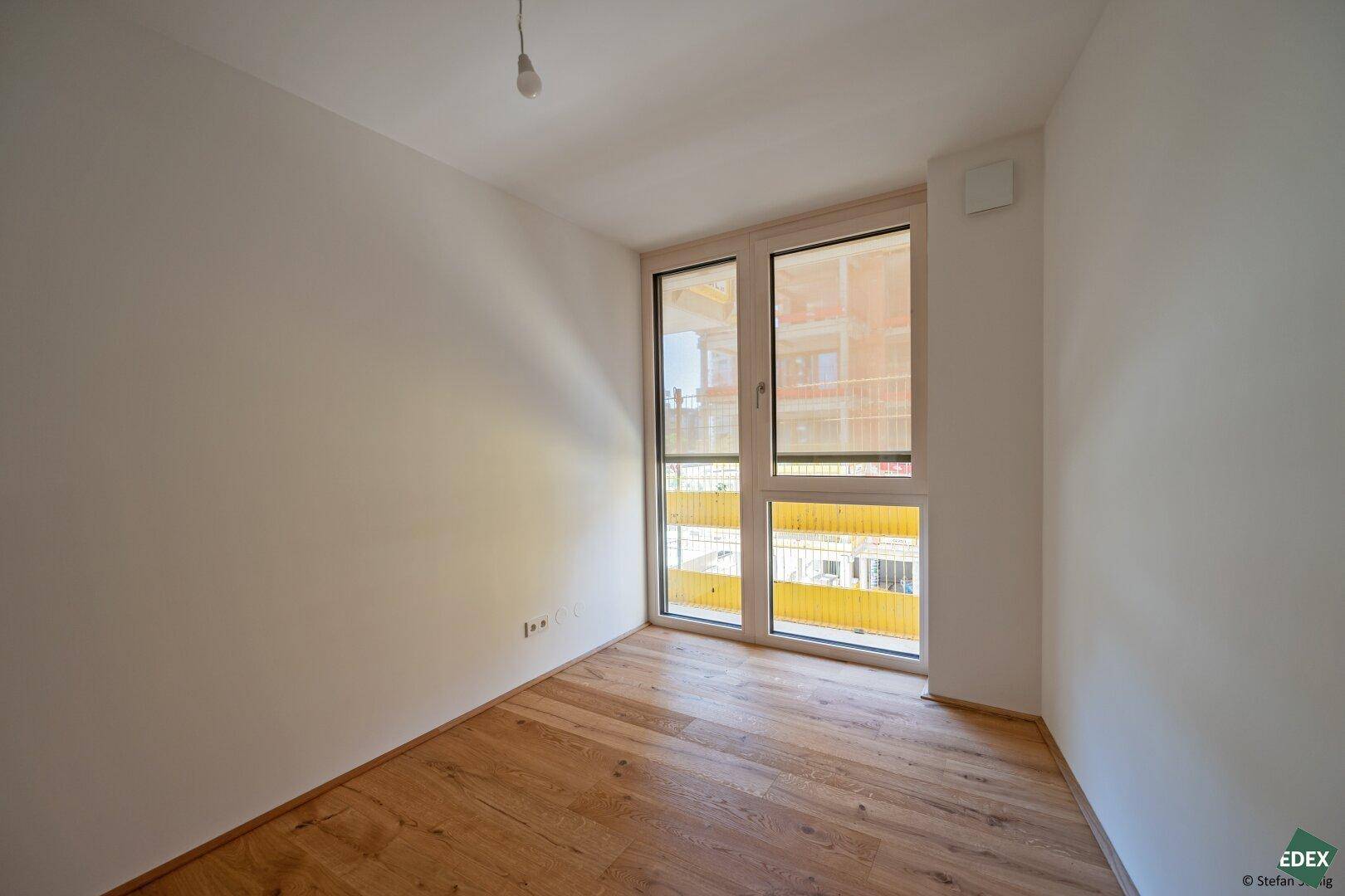 Helles Schlafzimmer mit Holzboden und großem Fenster, das viel Tageslicht hereinlässt.