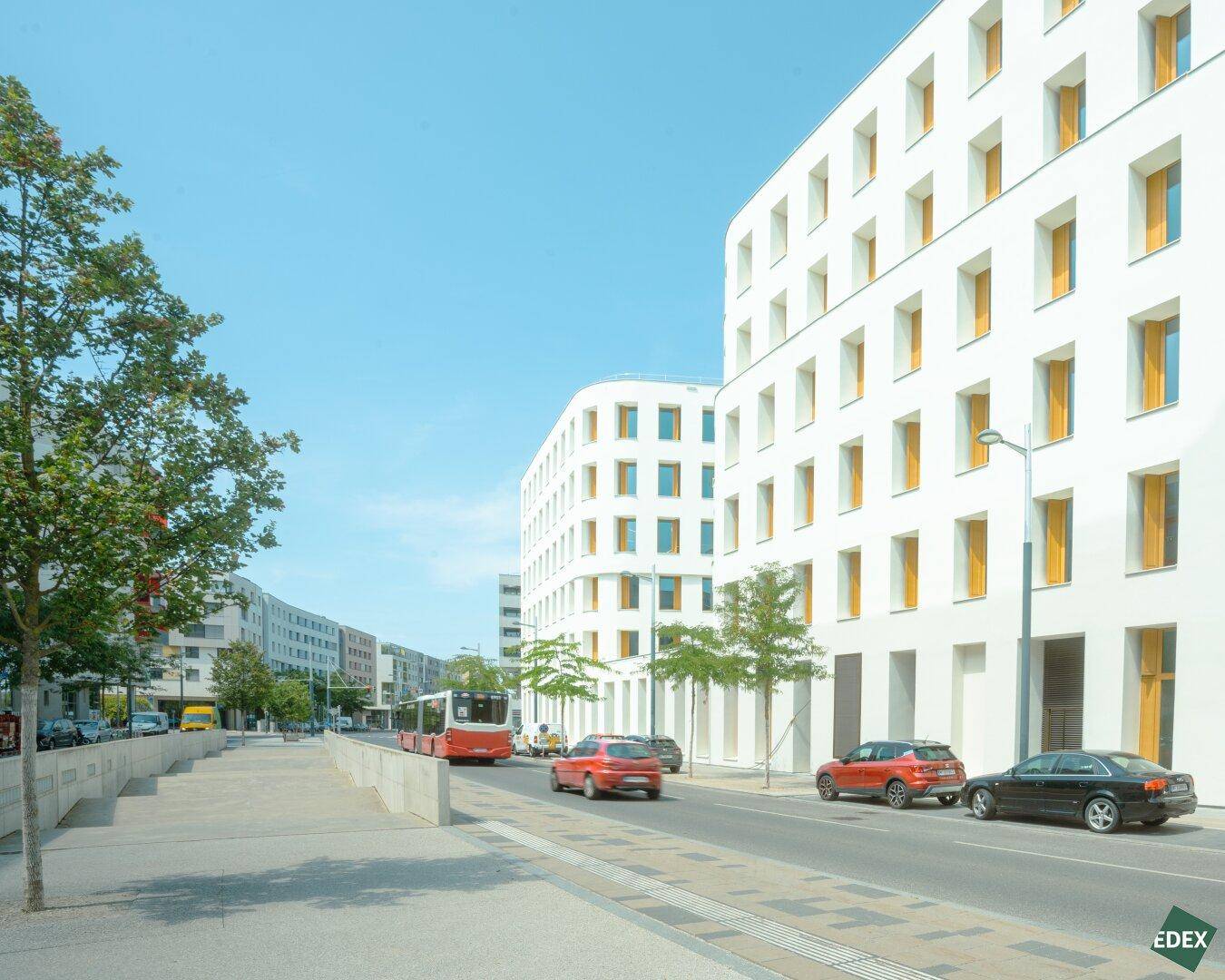 Moderne Gebäudefassade entlang einer Straße mit fahrenden Autos und einem Bus an einem sonnigen Tag.