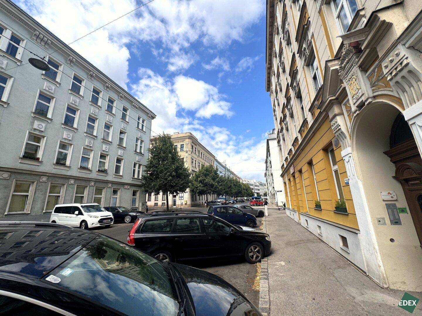 Straßenansicht mit Gebäuden, geparkten Autos und Bäumen unter blauem Himmel.