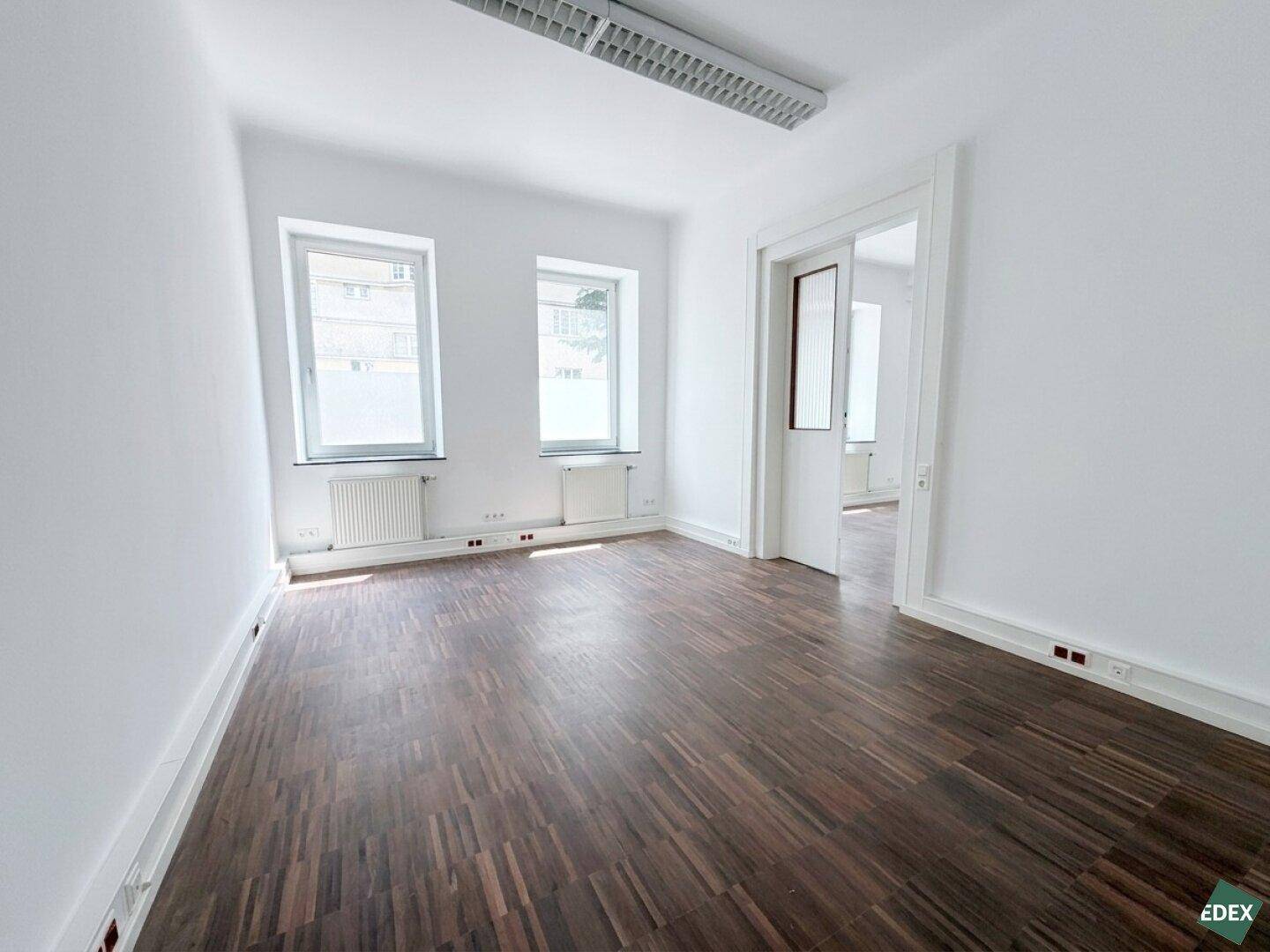 Geräumiger Büroraum mit Parkettboden und zwei Fenstern, durch eine Tür mit weiterem Bereich verbunden.