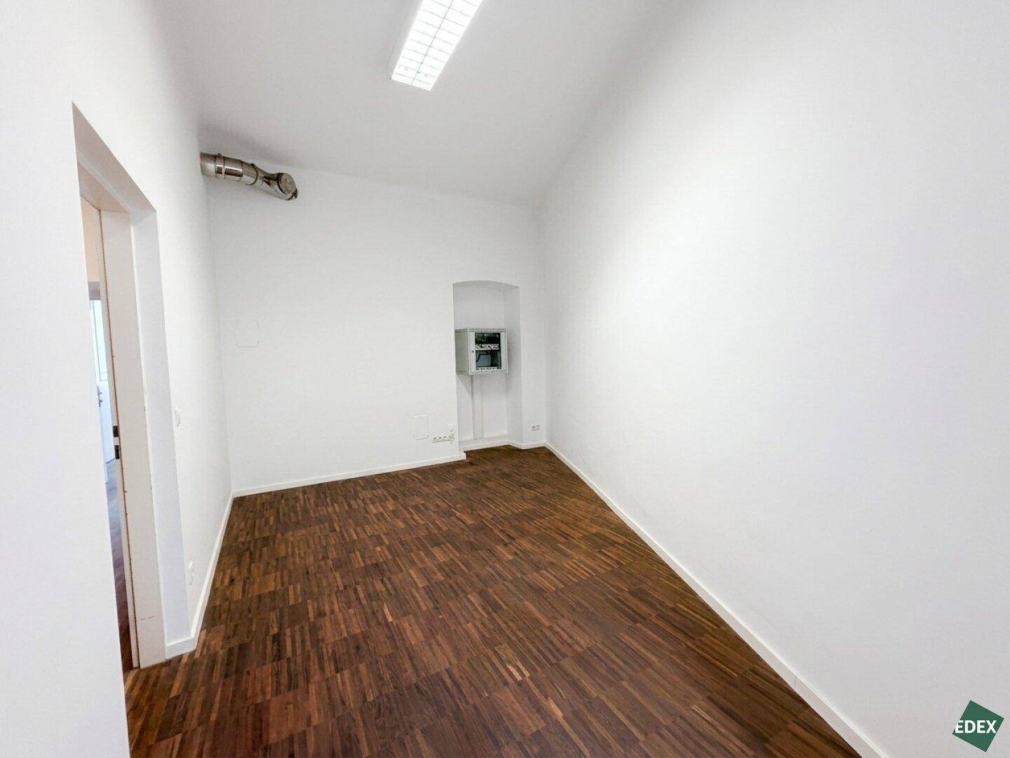 Kleiner, leerer Raum mit Parkettboden und sichtbaren Installationen an der Wand.