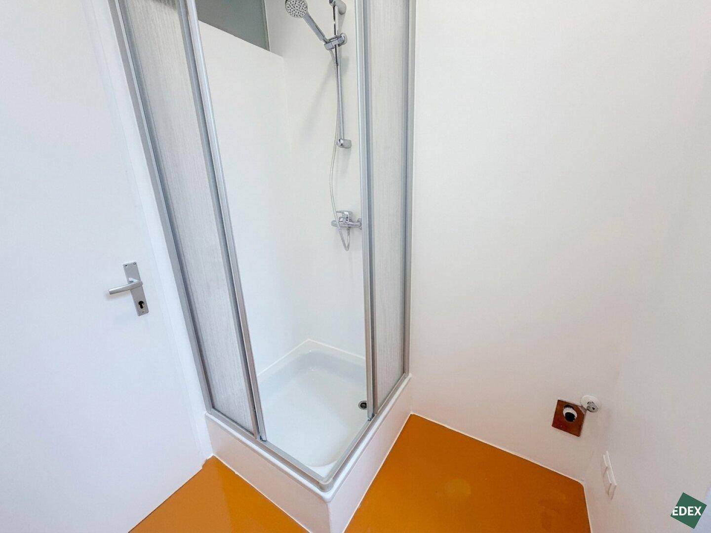 Duschbereich in einem hellen Badezimmer mit orangefarbenem Boden und weißer Duschkabine.