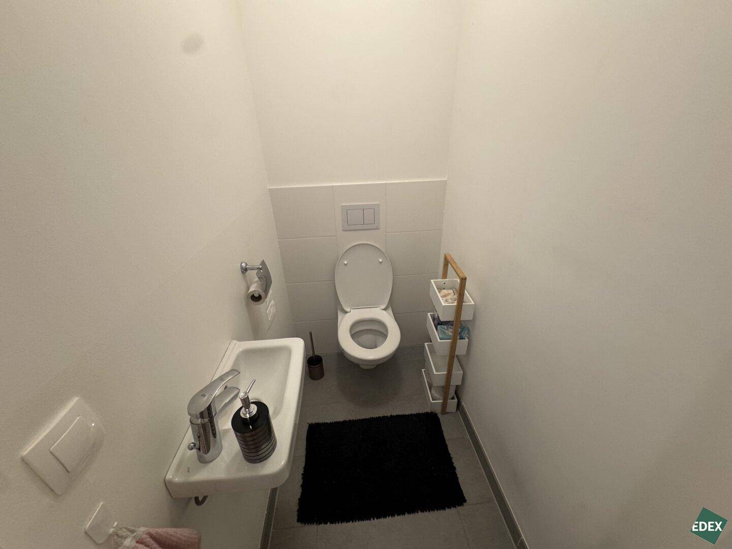 Kleines Gäste-WC mit Wand-WC, Handwaschbecken und einem schmalen Regal für Badutensilien.