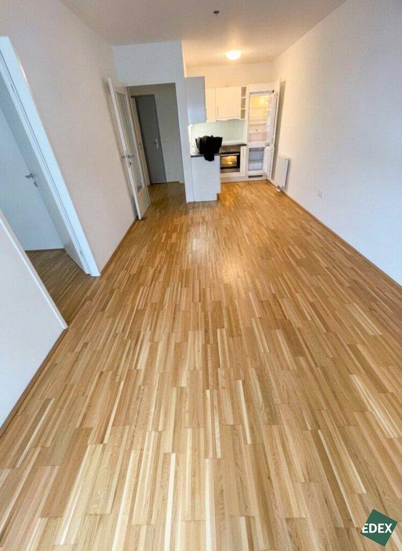 Offener Wohnbereich mit hellem Parkettboden, weißen Wänden und einer integrierten Küchenzeile im Hintergrund.