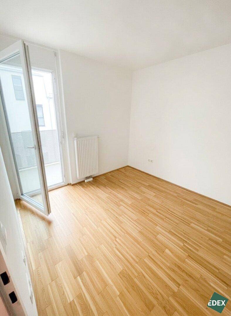 Helles Zimmer mit Parkettboden, weißen Wänden und Zugang zu einem Balkon, ideal als Schlafzimmer.