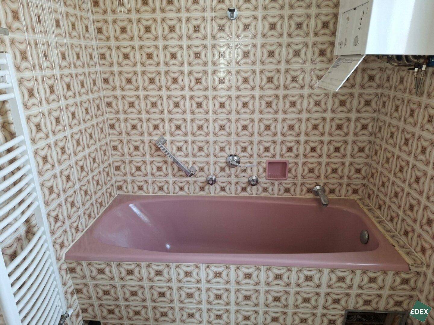 Badezimmer mit rosa Badewanne, gemusterten Fliesen und einem Handtuchheizkörper.