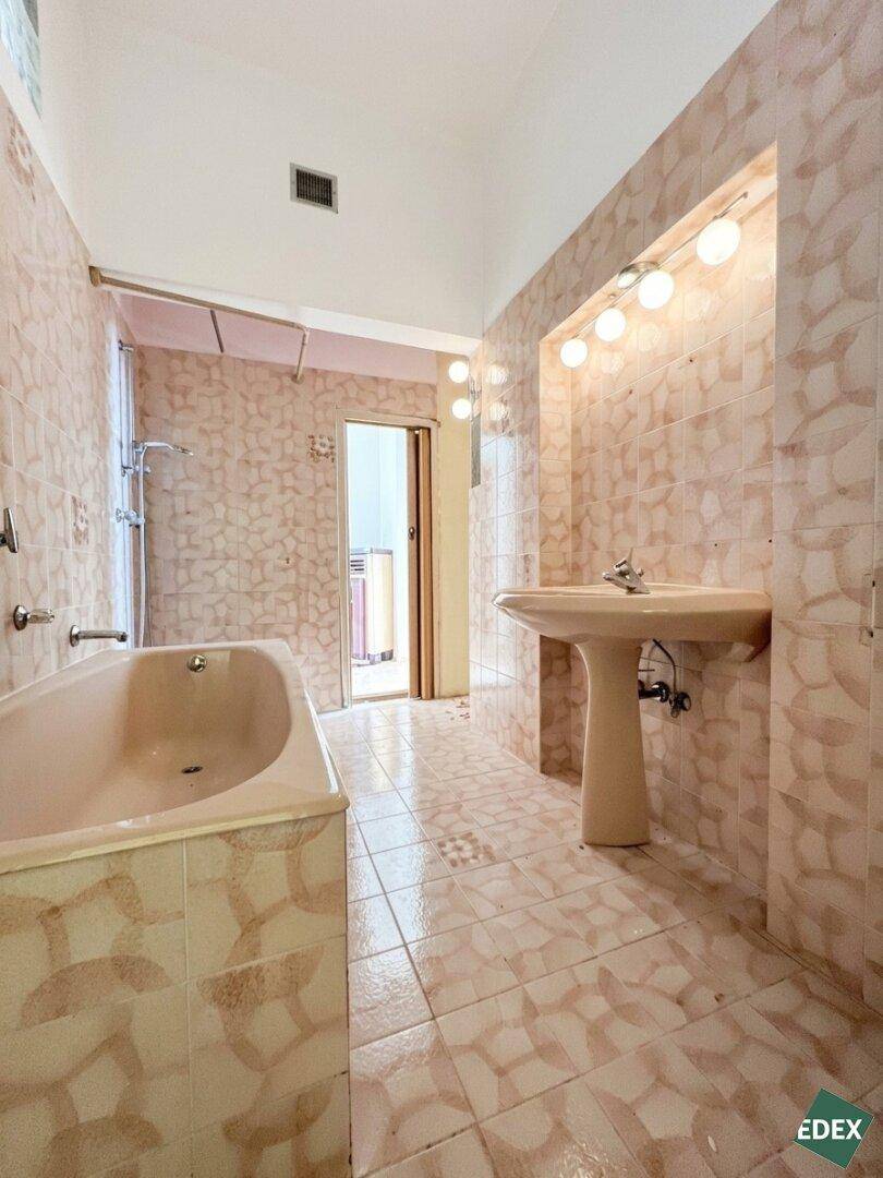 Veraltetes Badezimmer mit beigefarbenen Fliesen, Badewanne mit Duschfunktion und Waschbecken.