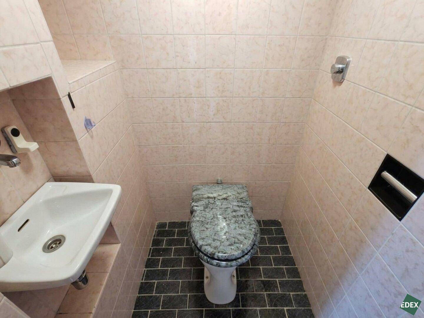 Separate Toilette mit rosa Wandfliesen, dunklem Boden und kleinem Handwaschbecken.
