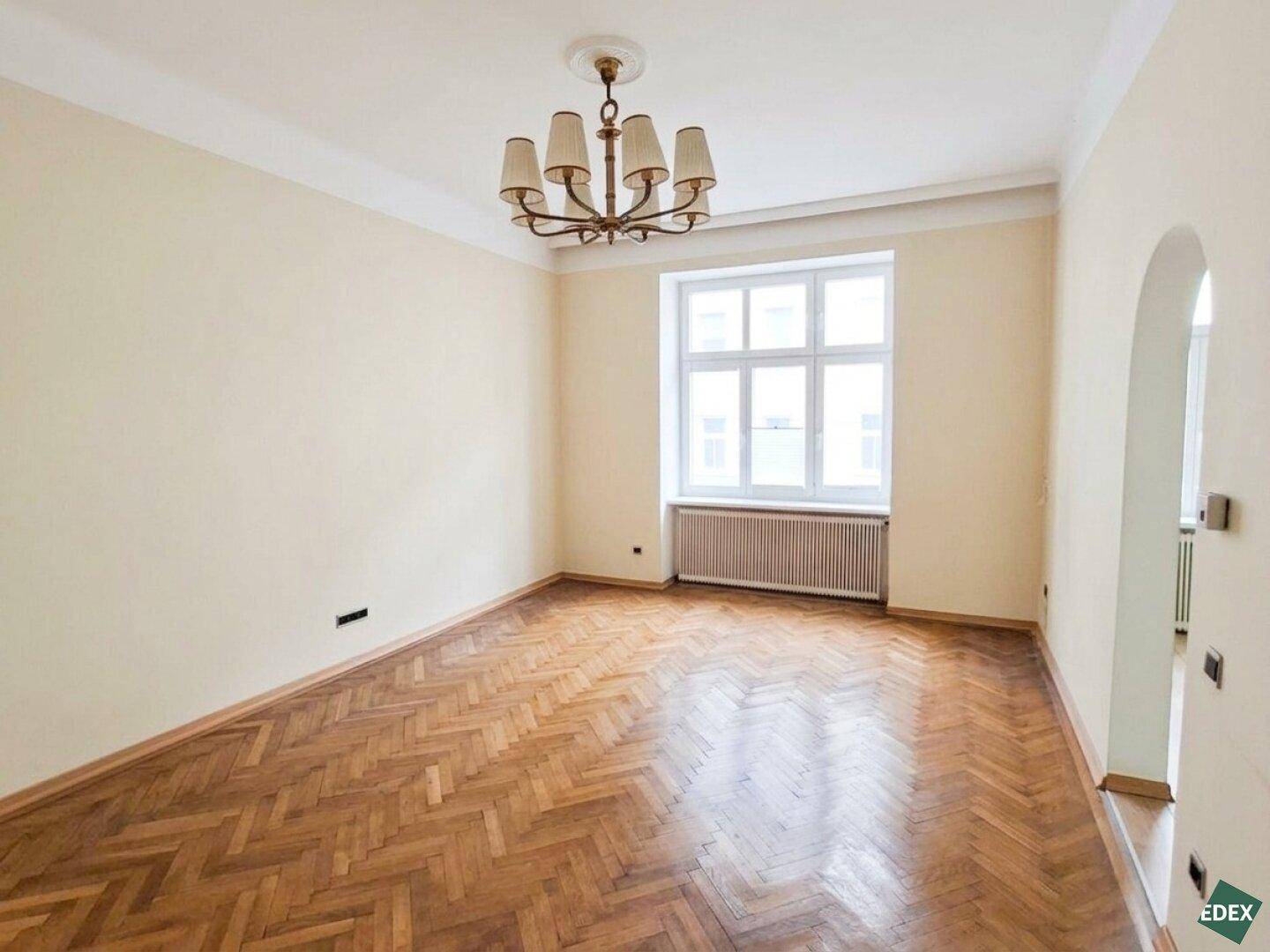 Geräumiges, leeres Zimmer mit Fischgrätparkett, hohen Decken und großem Fenster.