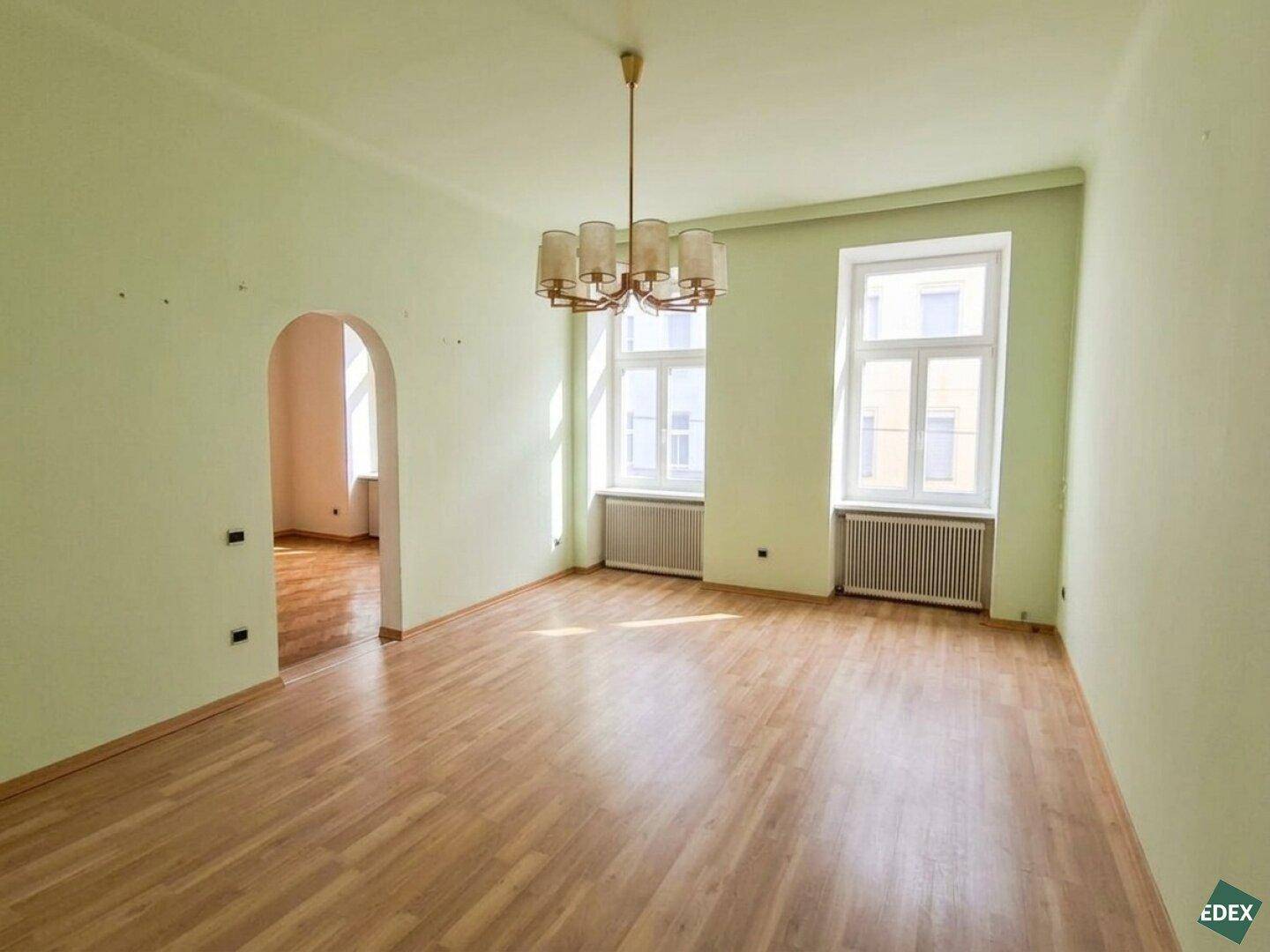 Großes, hellgrünes Zimmer mit Parkettboden, zwei Fenstern und einem klassischen Kronleuchter.