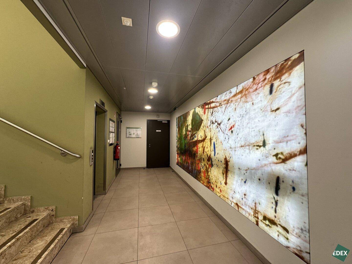 Eingangsbereich mit Treppe, Aufzug und einem großen, farbenfrohen Kunstwerk an der Wand.