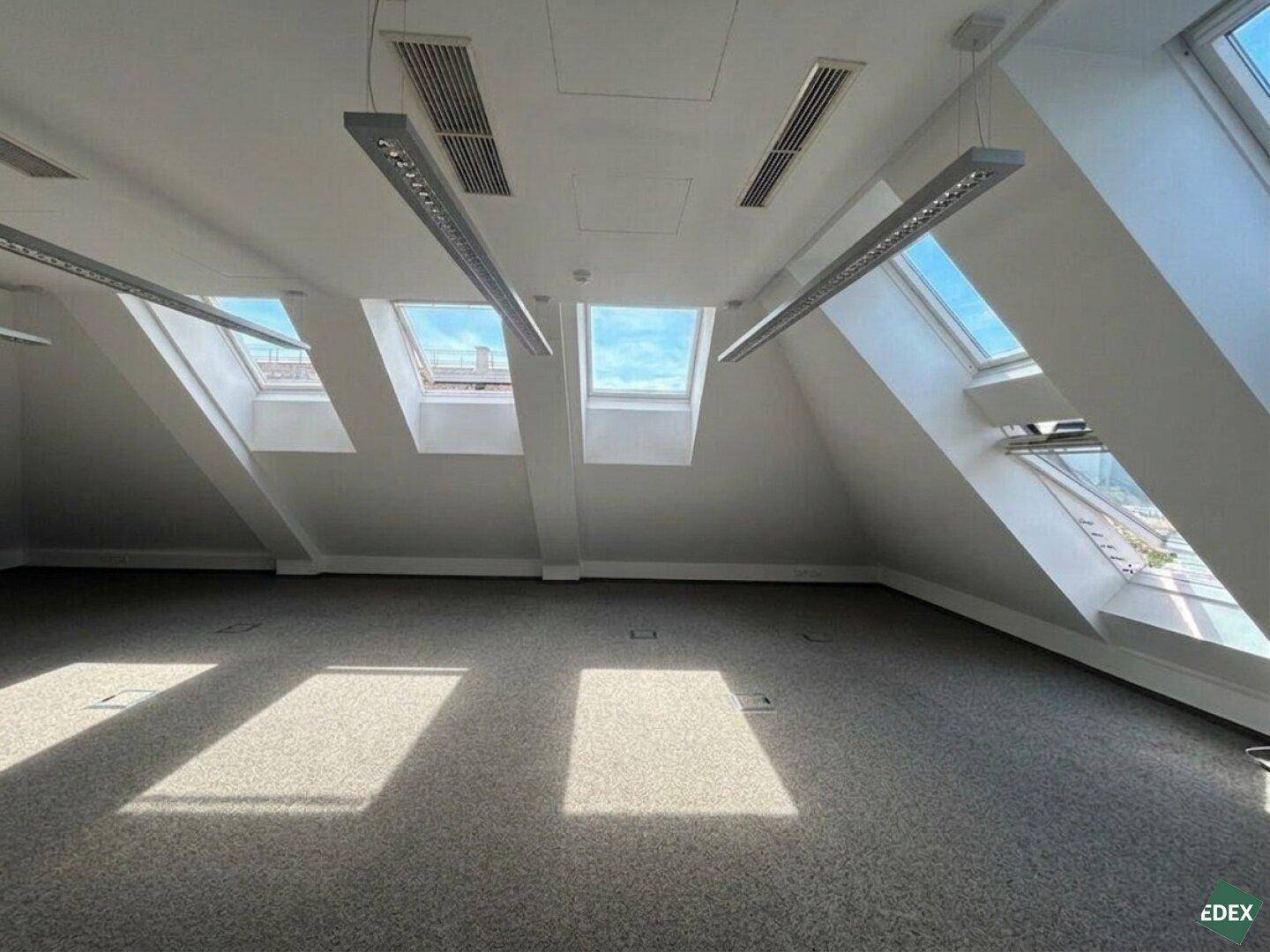 Geräumiges, leeres Büro im Dachgeschoss mit Teppichboden und viel Tageslicht durch Dachfenster.