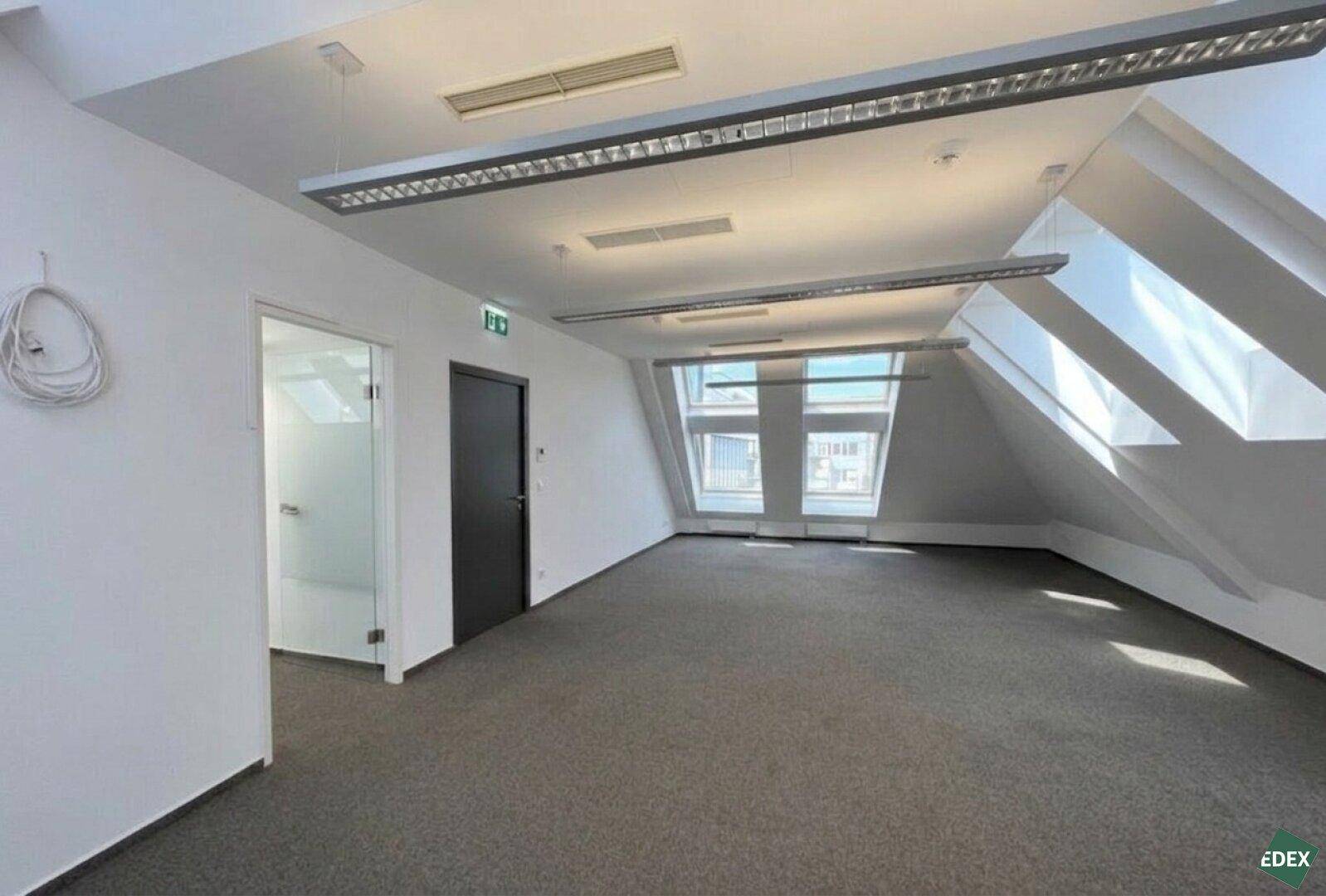 Leerer Büroraum im Dachgeschoss mit Teppichboden, Dachfenstern und Zugang zu einem weiteren Raum.