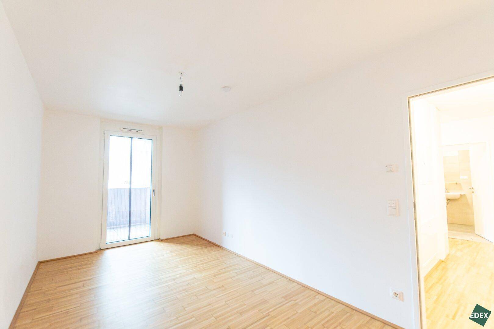 Helles Schlafzimmer mit Parkettboden und großem Fenster mit Zugang zum Balkon.