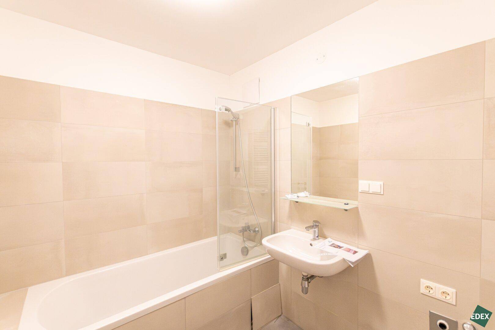 Modernes Badezimmer mit beigefarbenen Fliesen, Badewanne mit Duschwand und Waschbecken.