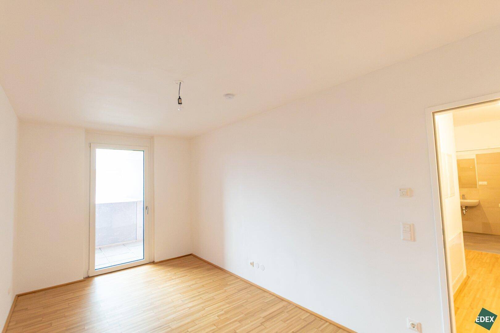 Helles Schlafzimmer mit Parkettboden und direktem Zugang zu einem Balkon, angrenzend an ein Badezimmer.
