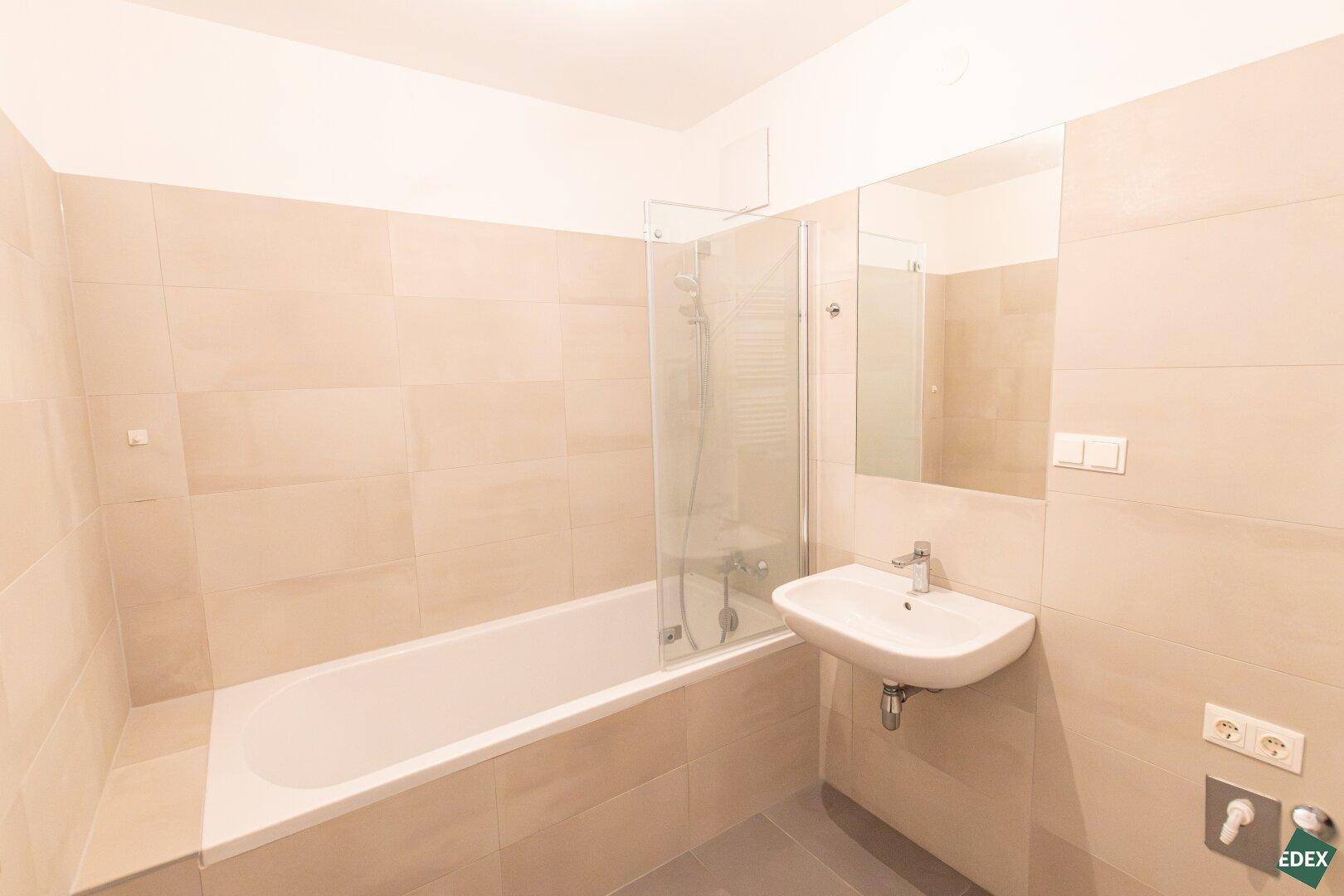 Modernes Badezimmer mit Badewanne, Duschwand, Waschbecken und beigefarbenen Fliesen.