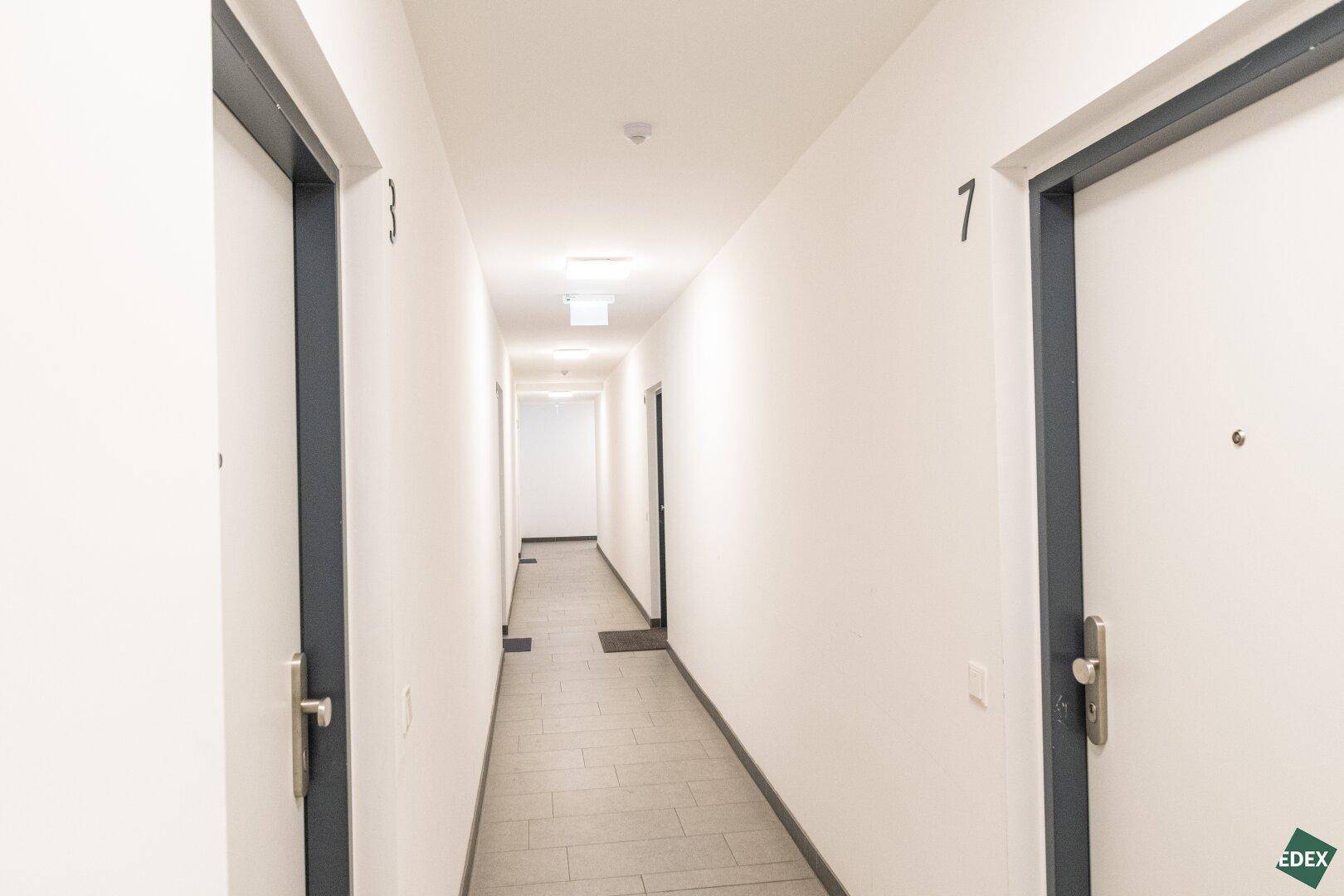 Langer, heller Flur im Gebäude mit mehreren Wohnungstüren und Fliesenboden.