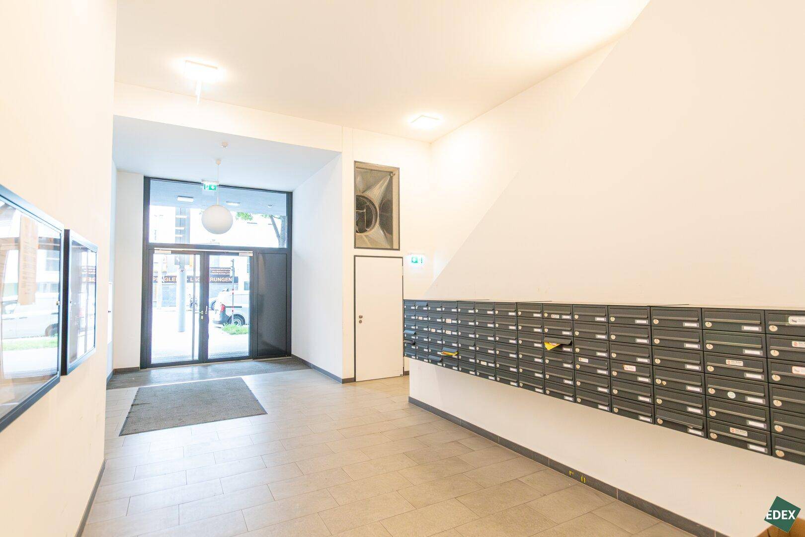 Heller Eingangsbereich des Gebäudes mit Briefkästen und großen Glastüren zur Straße.