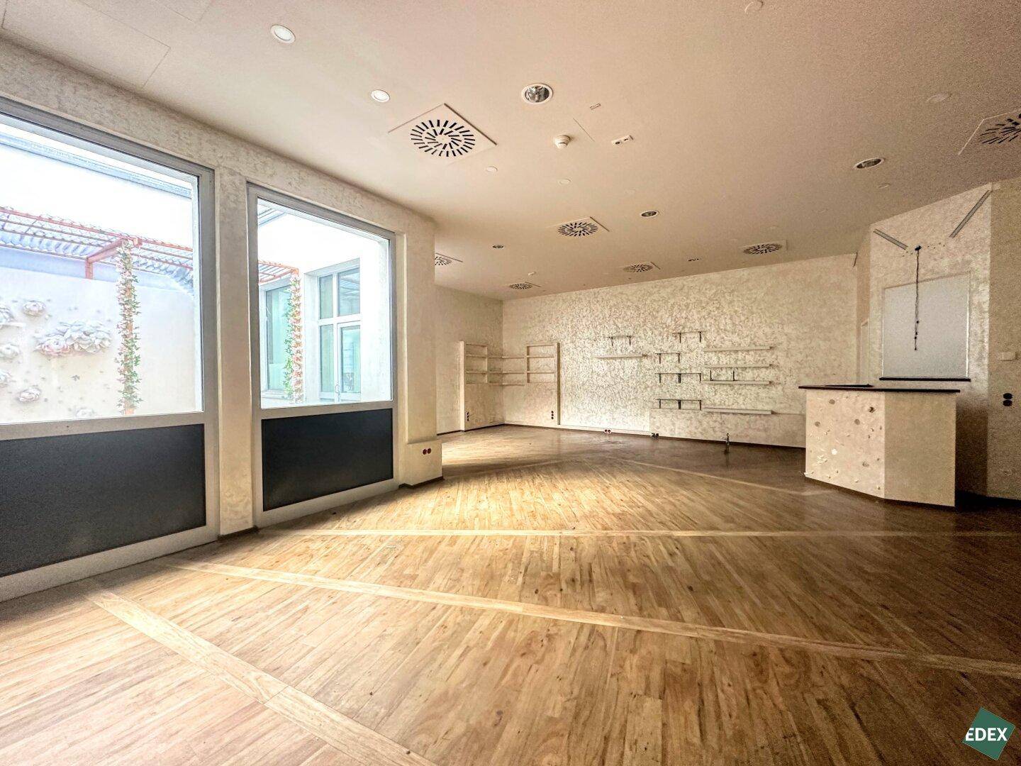 Heller Ladenraum mit großen Schaufenstern, Holzfußboden und strukturierten Wänden mit Blick auf einen Innenhof.