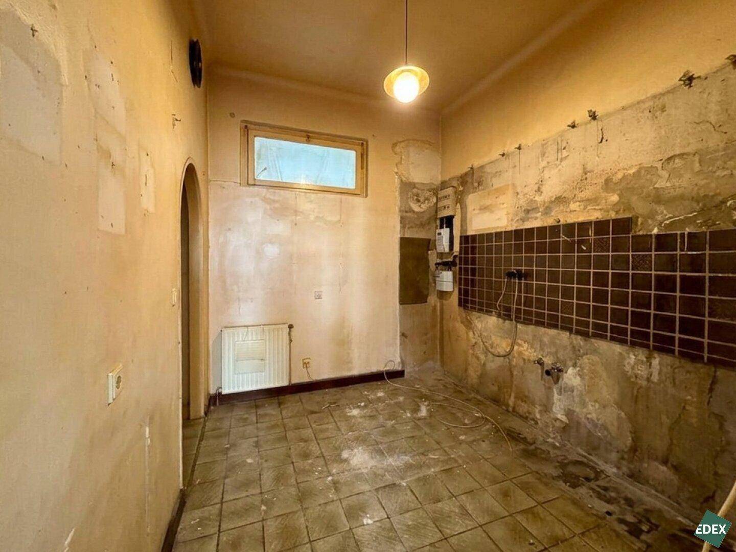 Sanierungsbedürftige Küche mit alten Fliesen, freiliegendem Putz und einem kleinen Fenster.
